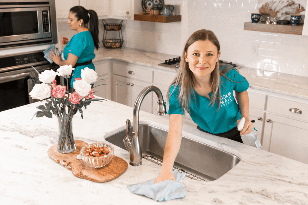 Two cleaners in teal uniforms tidy a bright white kitchen; one wipes the marble island at the sink, the other cleans near the oven in the background.