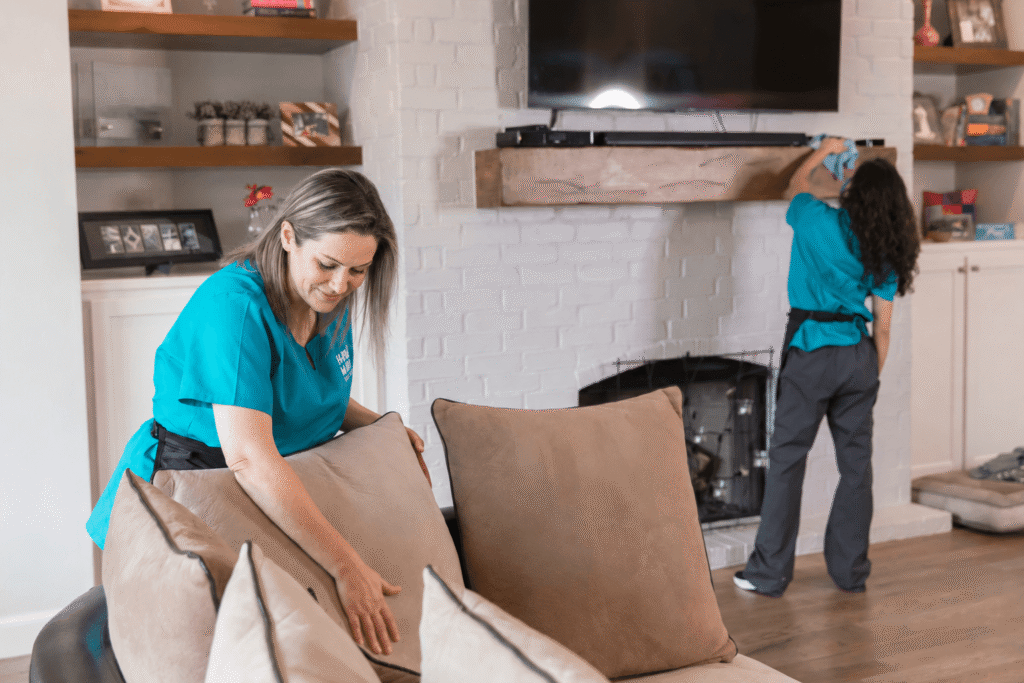 Two cleaners in teal uniforms rearranging beige cushions on a sofa in a living room while another cleaner wipes a shelf above a fireplace.